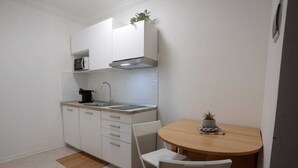 Een kleine keuken met witte kastjes, een magnetron, een fornuis en een spoelbak. Er staat een eettafel met twee stoelen en een bloempot erop.