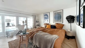 A modern bedroom with a large bed, a desk, and a chair. There are framed artworks on the walls and a window with curtains.