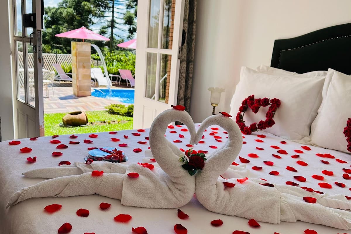 A bed with rose petals and a heart-shaped towel arrangement, a view of a swimming pool, and a patio area.