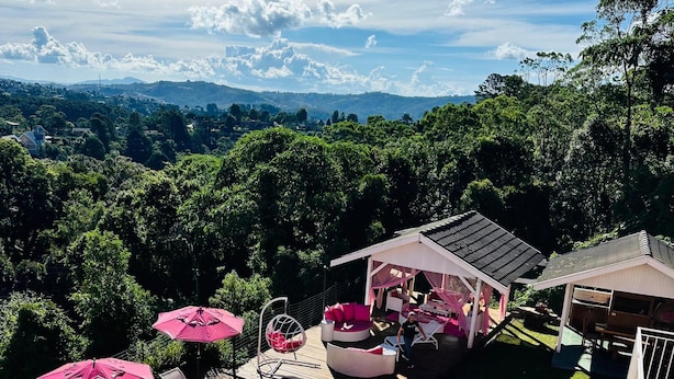A swimming pool with lounge chairs and pink umbrellas, surrounded by a deck and a view of a forested landscape. - Pousada Pink Village  (Campos do Jordão)