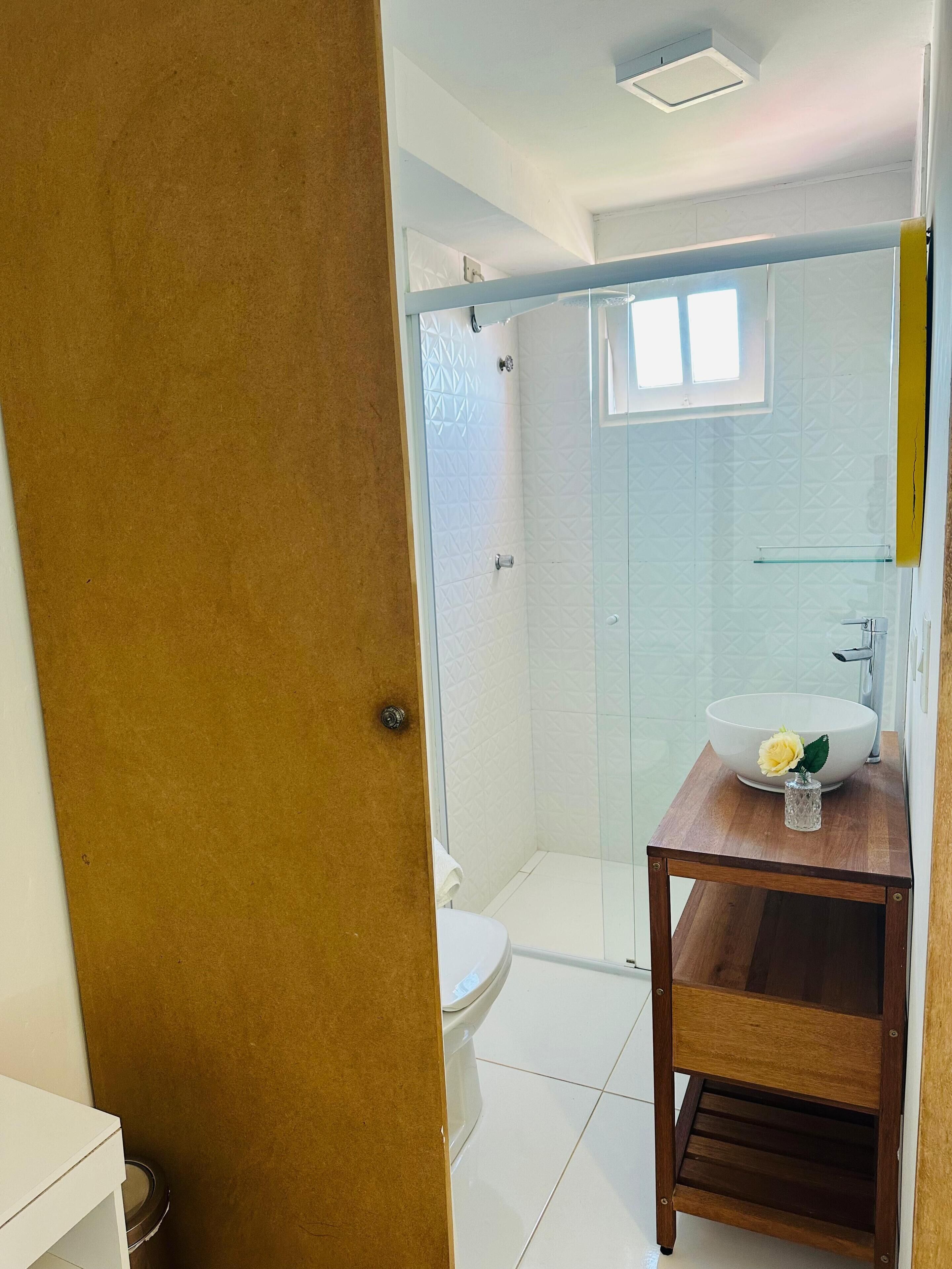 A bathroom with a white toilet, a glass-enclosed shower, and a wooden vanity with a white sink and a vase with a yellow flower.