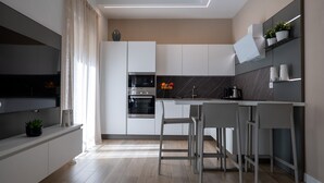 A modern kitchen with a built-in oven, a dining area with bar stools, and a large window with curtains. - Appartamento bilocale ARCADIA (Verona)