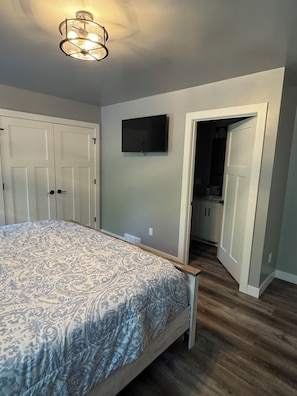 A bedroom with a bed, a flat-screen TV mounted on the wall, and a hallway leading to a bathroom. - Heart of Door County Home-Sleeps 16+ w/Hot Tub (Egg Harbor)