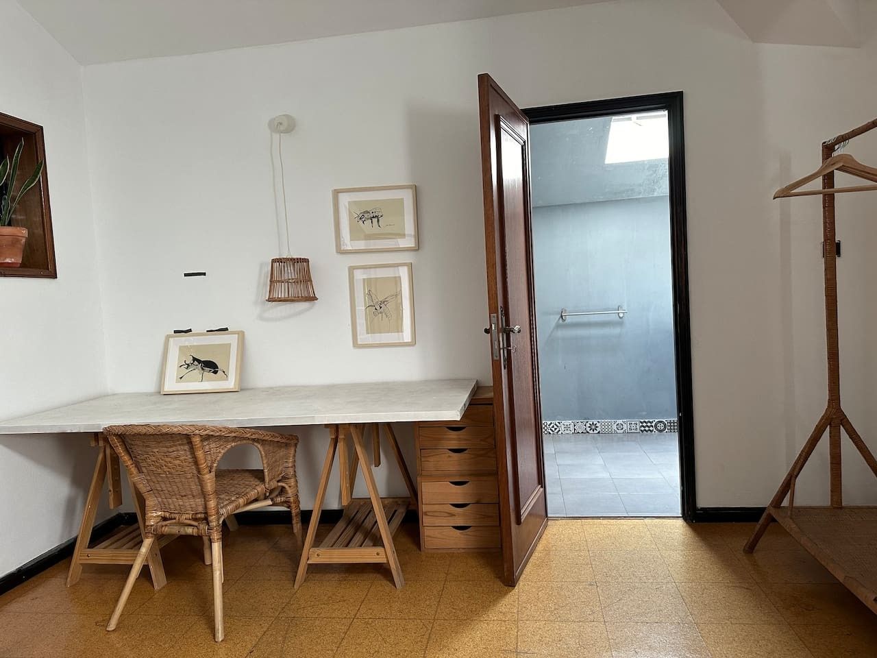 Une pièce avec un bureau blanc, des chaises en osier, une armoire en bois et une porte donnant sur un balcon.