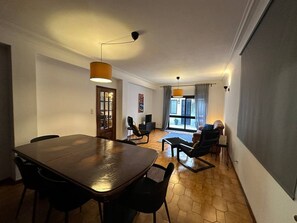 Un comedor con una gran mesa de madera y sillas, un sofá y una mesa baja. Hay una puerta que conduce a otra habitación y una ventana con cortinas.