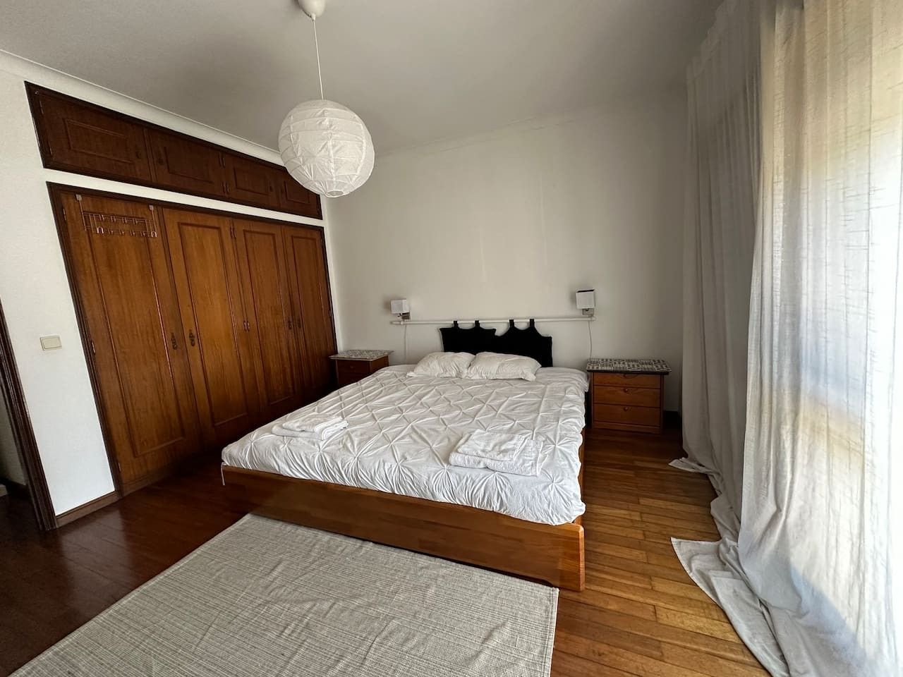 A bedroom with a large bed, wooden wardrobe, bedside tables, and a hanging light fixture.