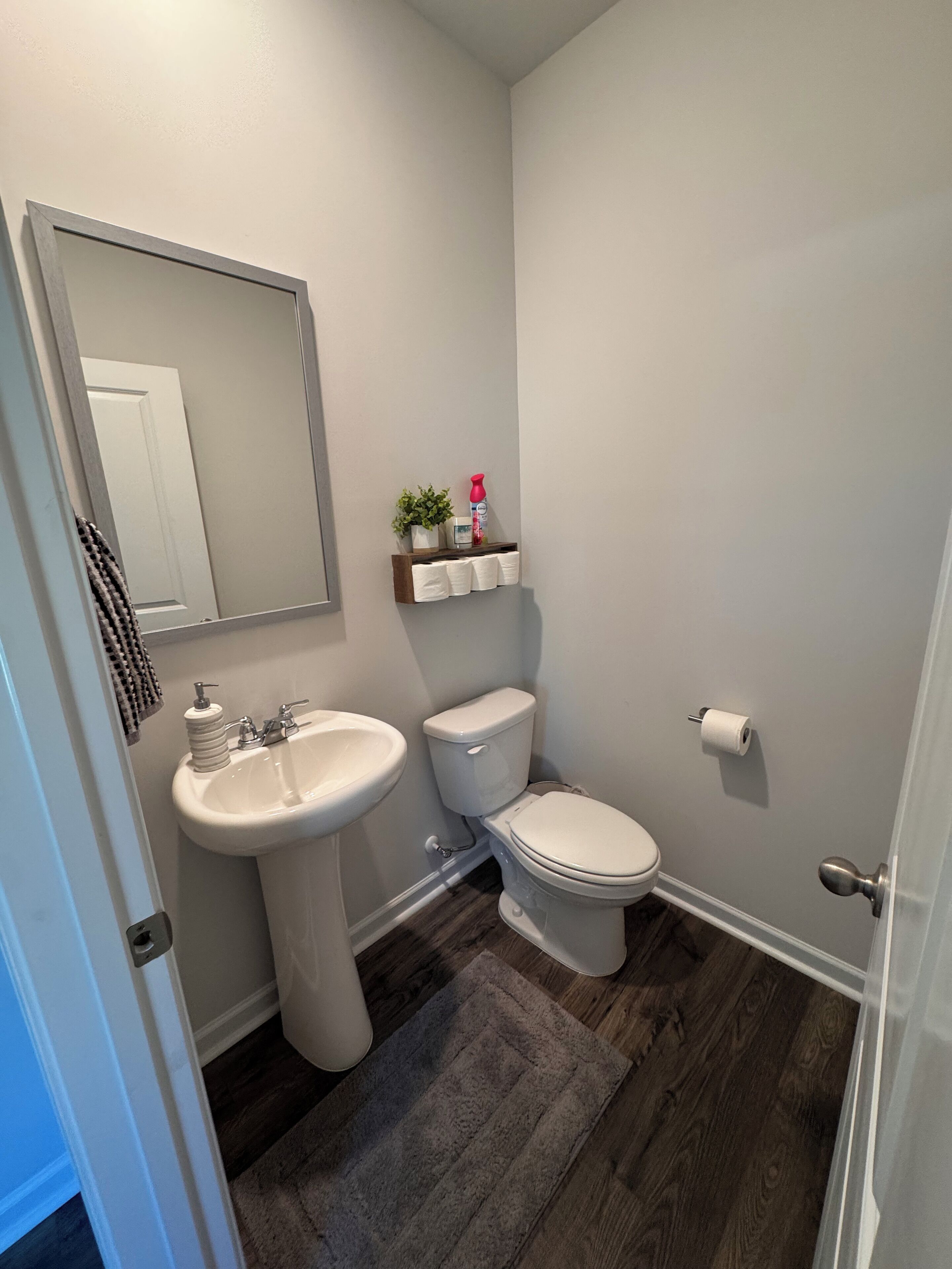 A bathroom with a pedestal sink, toilet, and a mirror. There is a towel rack with a towel and a small shelf with toiletries.