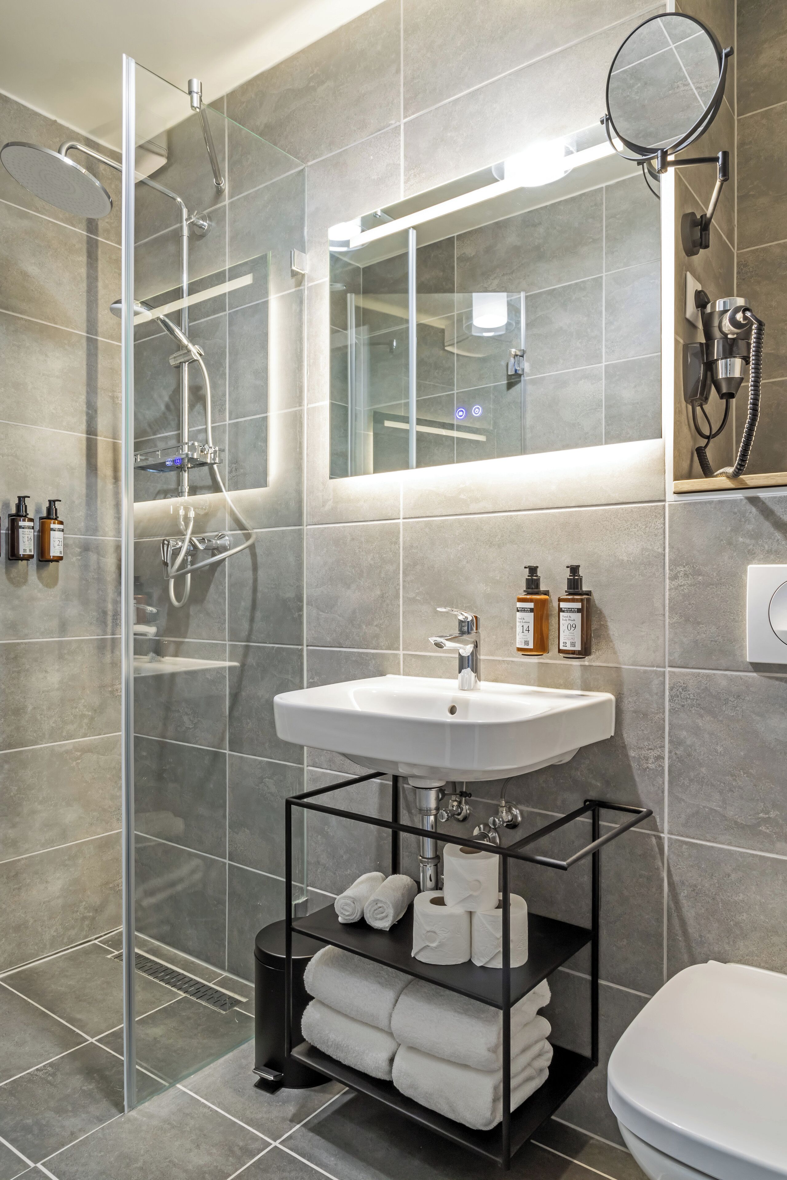 Un baño moderno con ducha de vidrio, un lavabo blanco y un espejo encima. Se ven dos botellas de champú y un toallero con toallas.