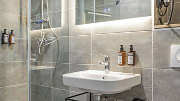 Un baño moderno con ducha de vidrio, un lavabo blanco y un espejo encima. Se ven dos botellas de champú y un toallero con toallas.