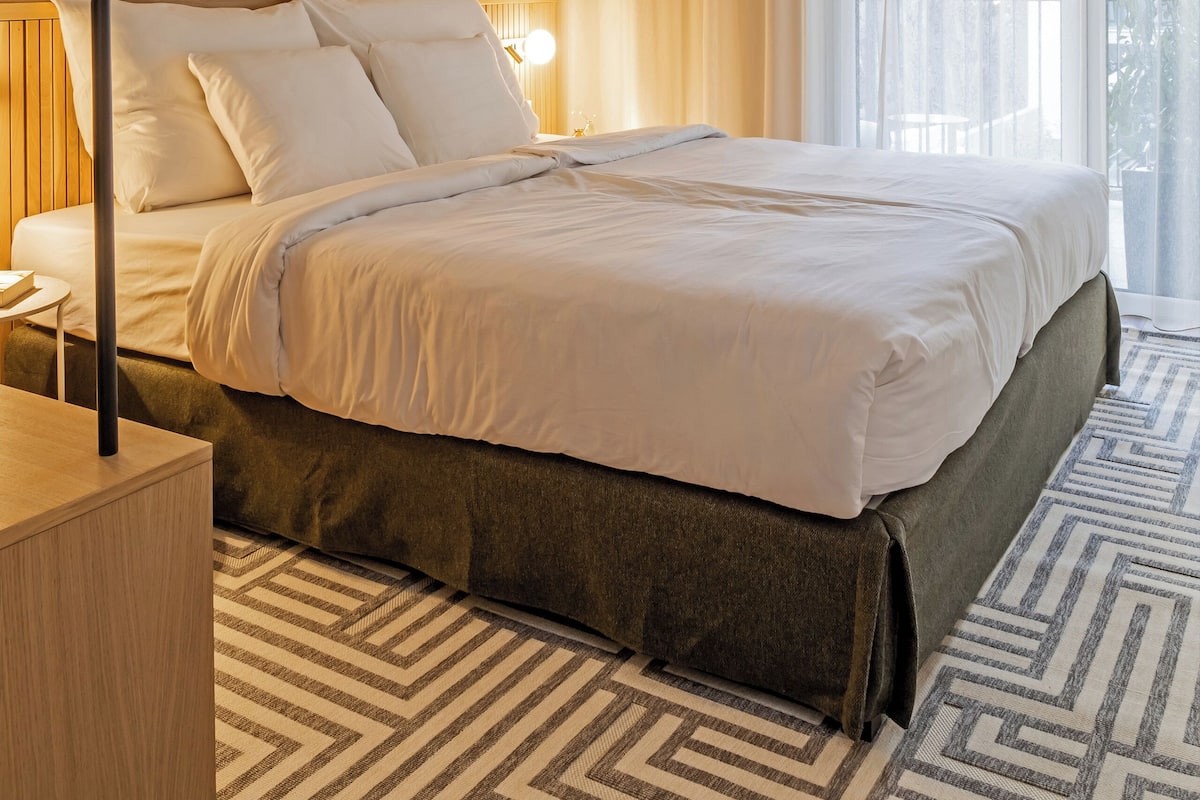 A modern hotel room with a large bed, wooden paneling, a geometric patterned rug, and a large window with curtains.