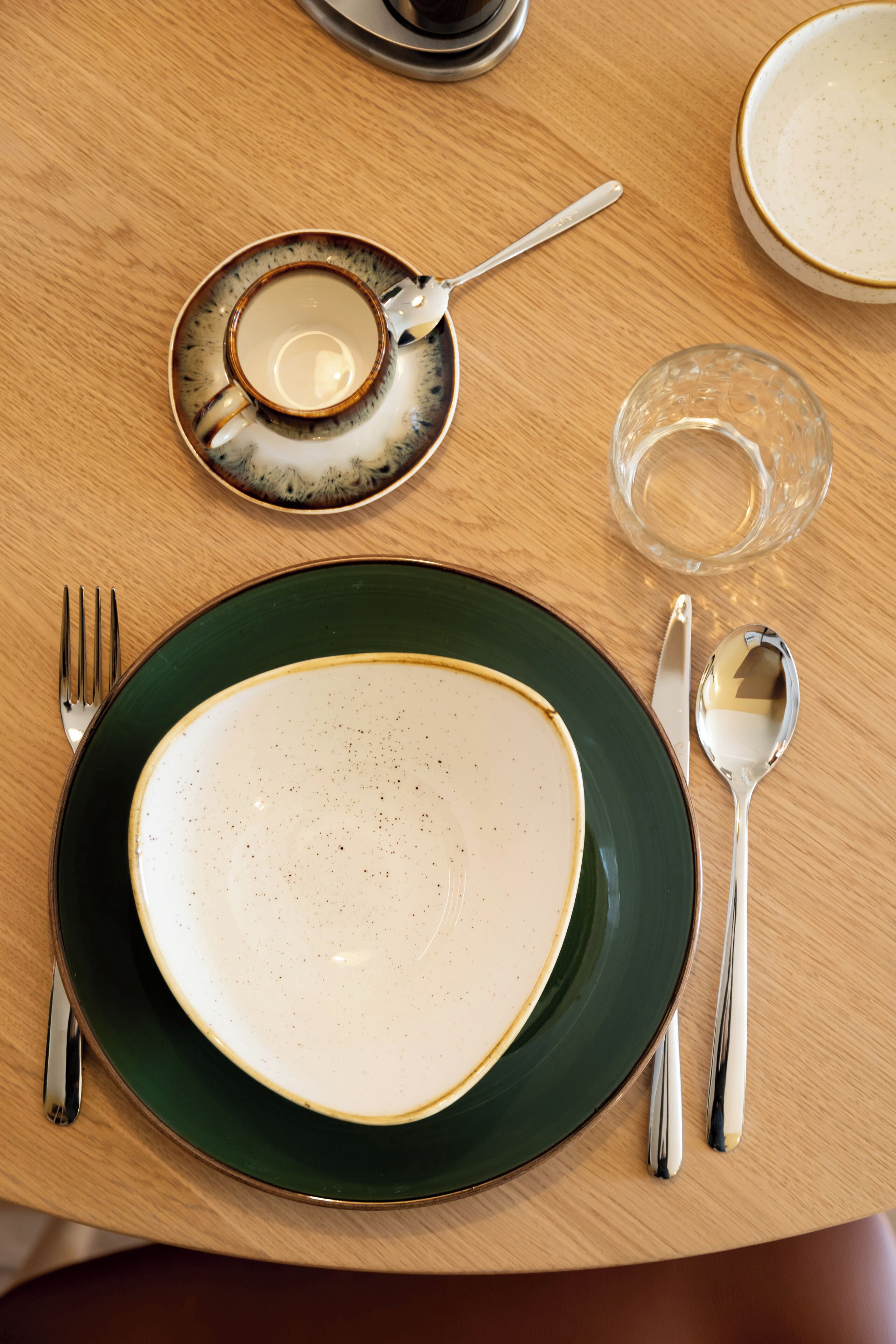 Una mesa dispuesta con un plato verde, una copa de cristal transparente, una taza y cubiertos.