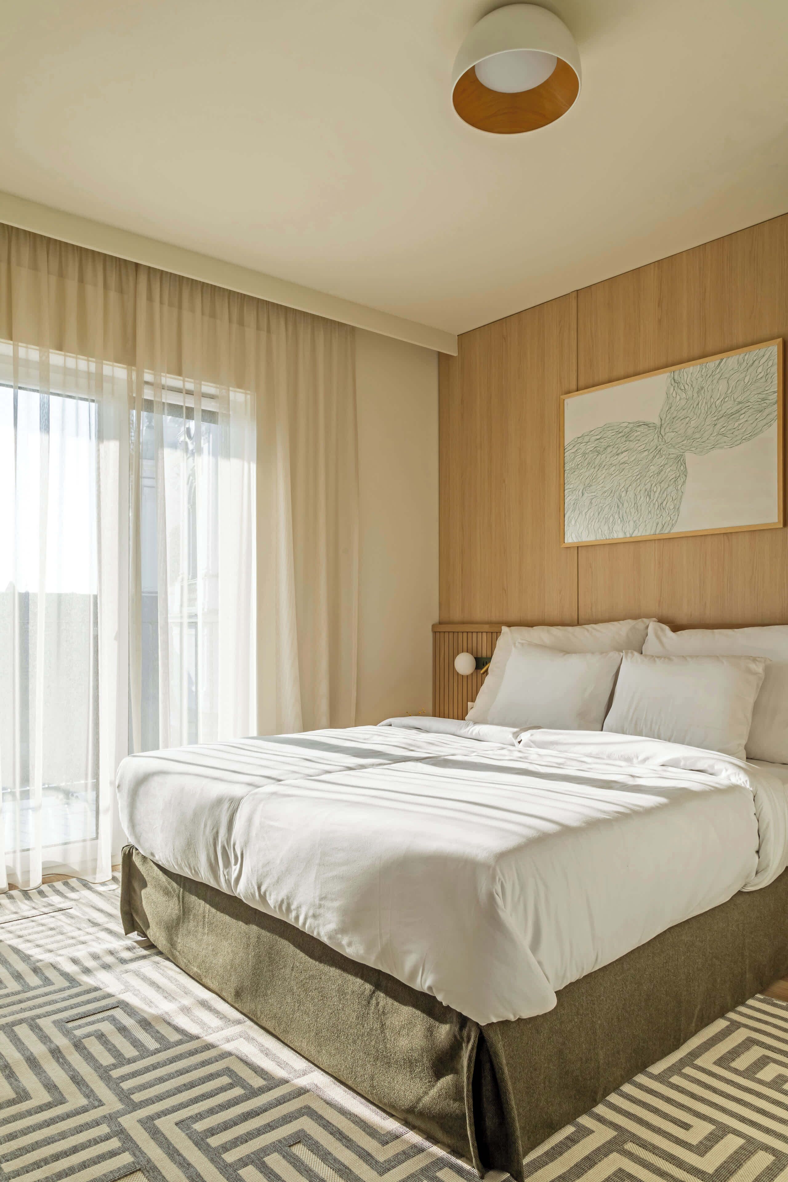 Une chambre d’hôtel avec un grand lit, une tête de lit en bois, une œuvre d’art accrochée au mur et une fenêtre ornée de rideaux légers.