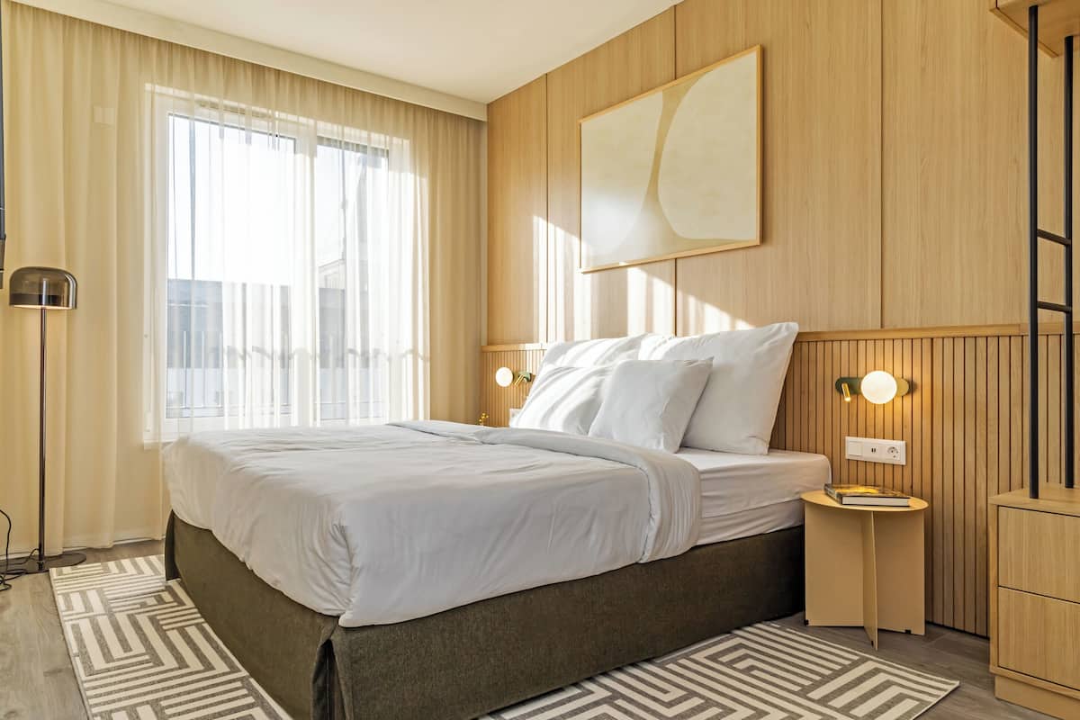 A hotel room with a large bed, a bedside table with a lamp, a wall-mounted artwork, and a view of the city through the window.