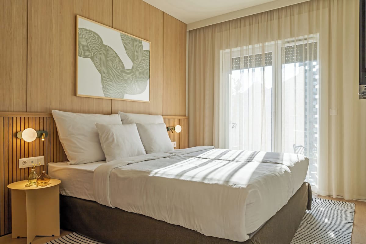 A modern hotel room with a large bed, wooden panel walls, a framed artwork, two bedside lamps, and a large window with curtains.
