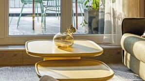 A modern living room with a large window, a sofa, a small table with a vase, and a view of the outside balcony. - Marone Suites Budapest (Budapest)
