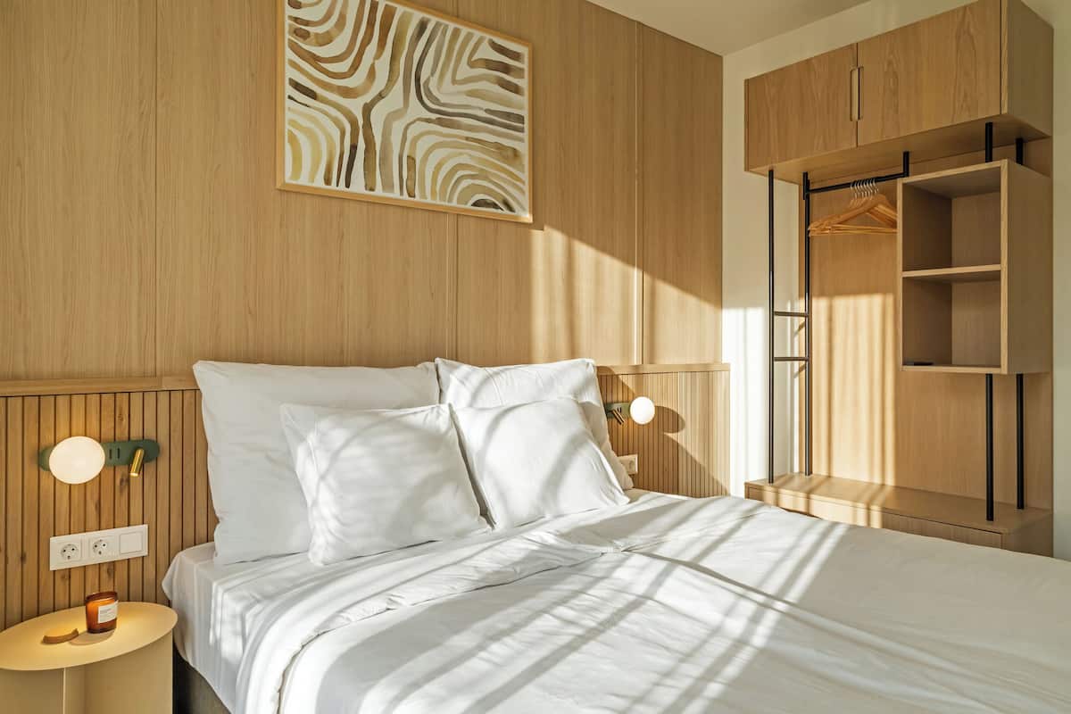 A modern bedroom with a wooden headboard, a bedside table with a lamp, and a wardrobe with a hanging rod.