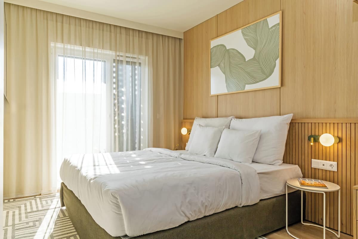 A hotel room with a large bed, a small round table, a wall-mounted light, and a framed artwork.