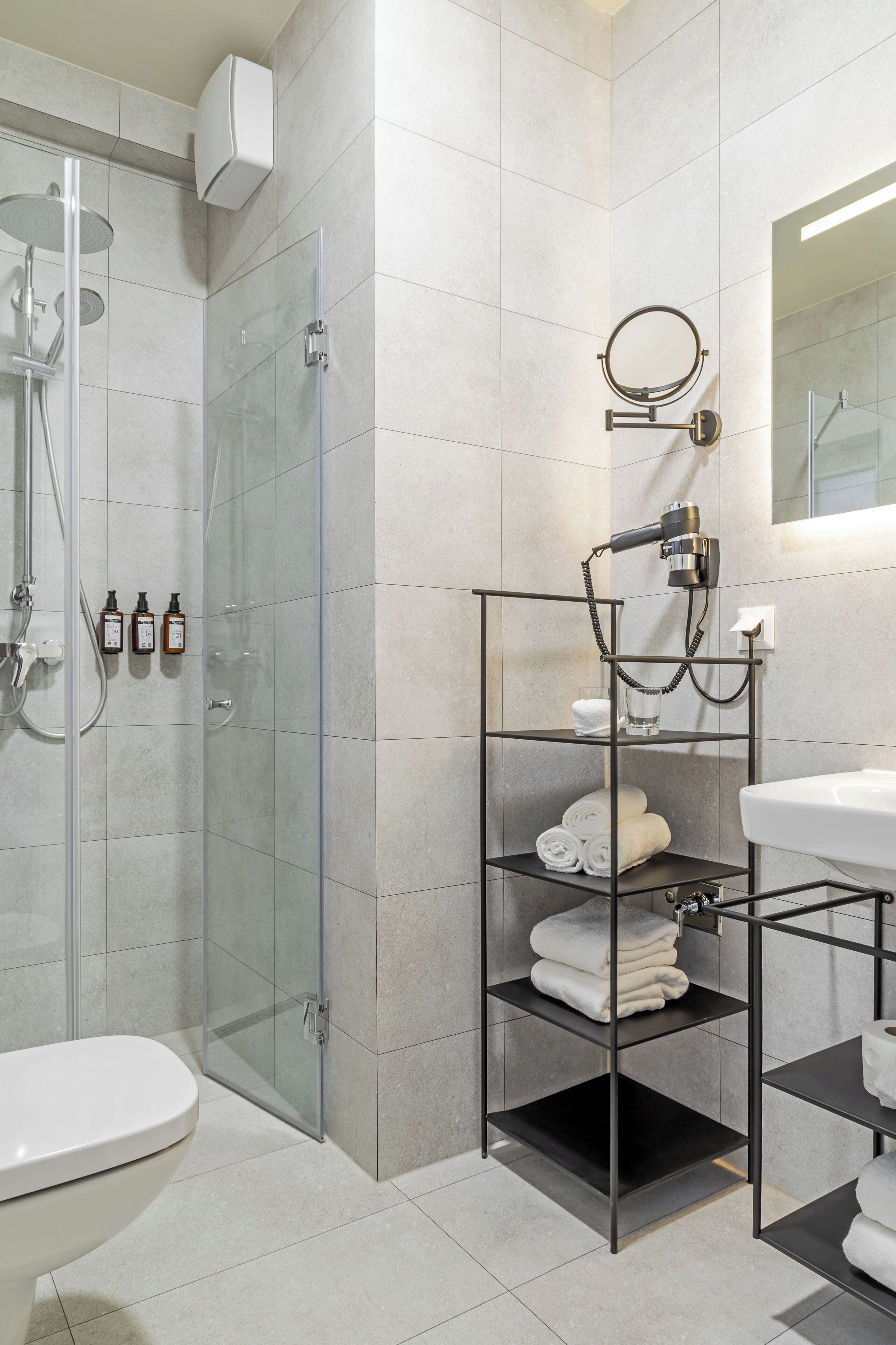 A modern bathroom with a glass-enclosed shower, a toilet, and a sink with a mirror. There are shelves with towels and a hairdryer.