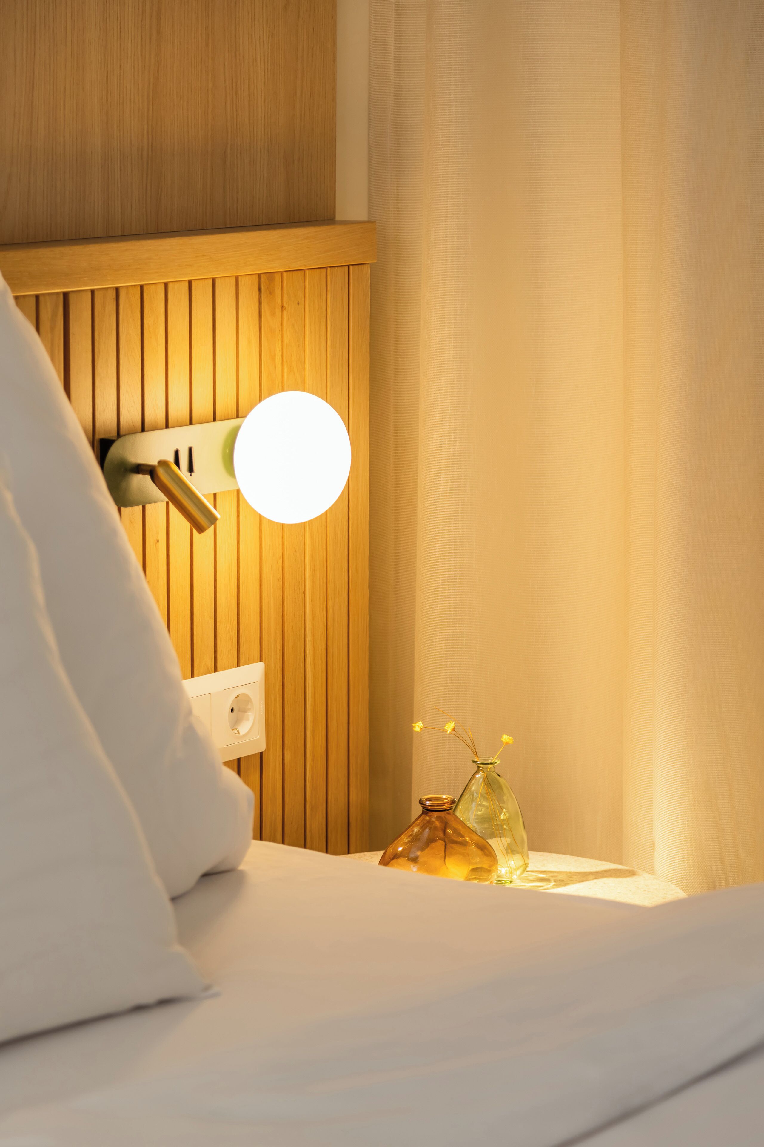 A neatly made bed with white bedding, a wooden headboard, a wall-mounted round mirror, and a small vase with flowers on a bedside table.