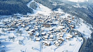 Uma aldeia coberta de neve com edifícios agrupados, uma pista de esqui e montanhas ao fundo.