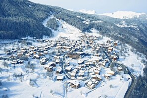 Ein verschneites Dorf mit zusammengeklammerten Häusern, einer Skipiste und Bergen im Hintergrund.