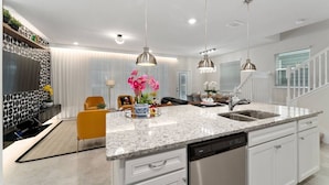 A modern kitchen with granite countertops, white cabinetry, and stainless steel appliances. There are pendant lights, a built-in oven, and a large island with a sink. - Themed 5BR Home w/ Pool · Windsor Island Resort (Davenport)