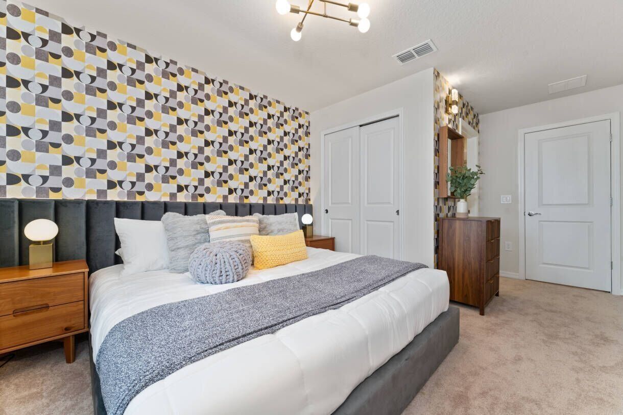 A bedroom with a geometric patterned wall, a bed with multiple pillows, a bedside table with a lamp, and a dresser.