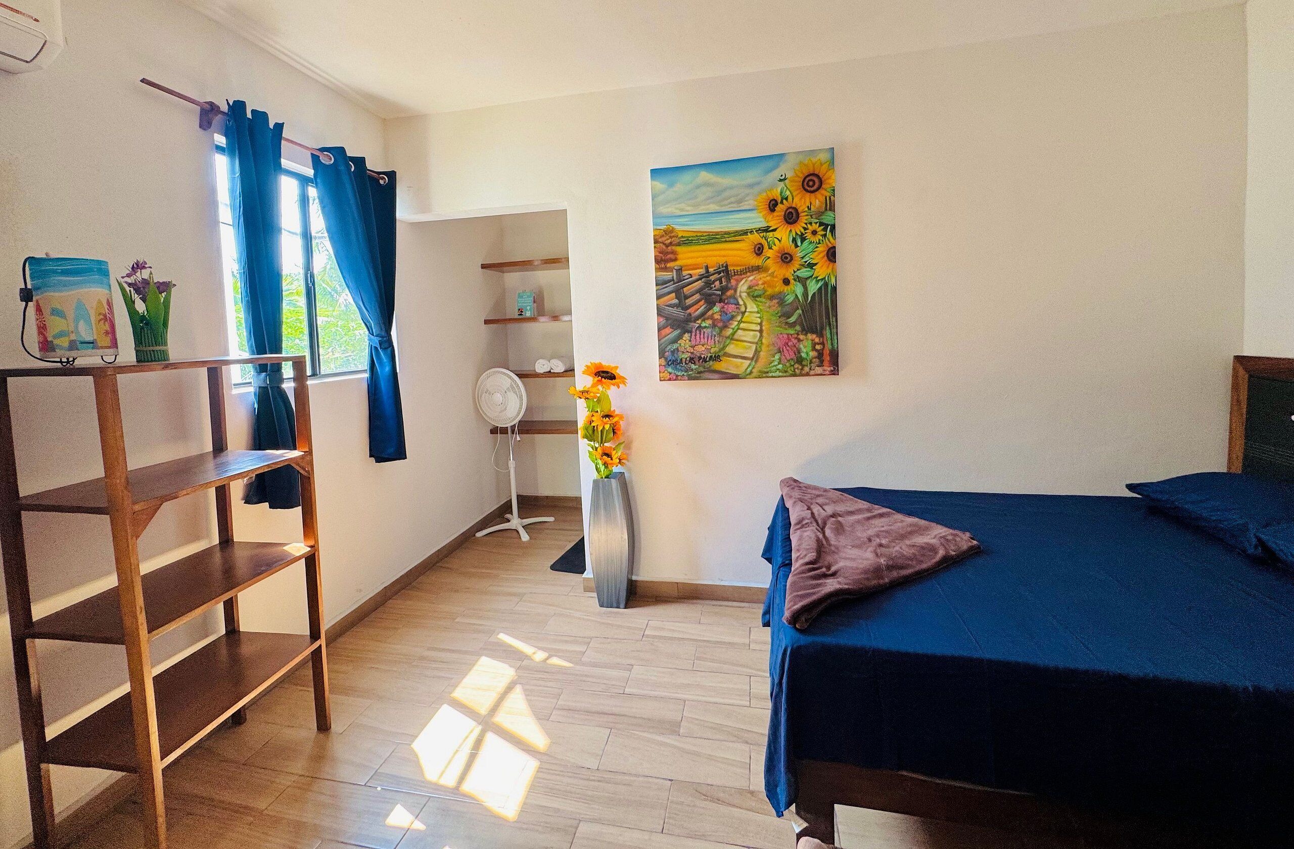 A bedroom with a bed, a wooden shelf, a fan, a vase with flowers, and a painting of sunflowers.