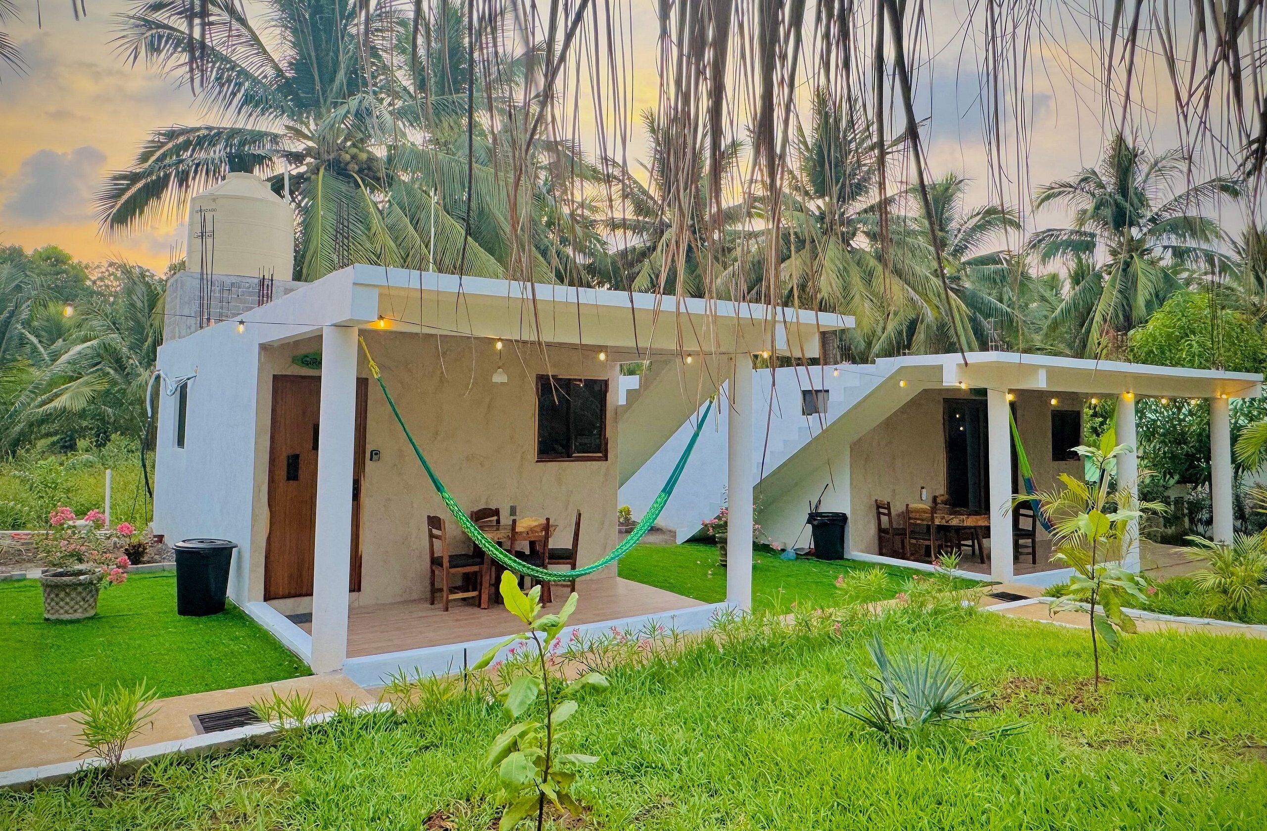 Een modern huis met twee verdiepingen en een overdekte patio, omgeven door weelderige begroeiing en palmbomen.