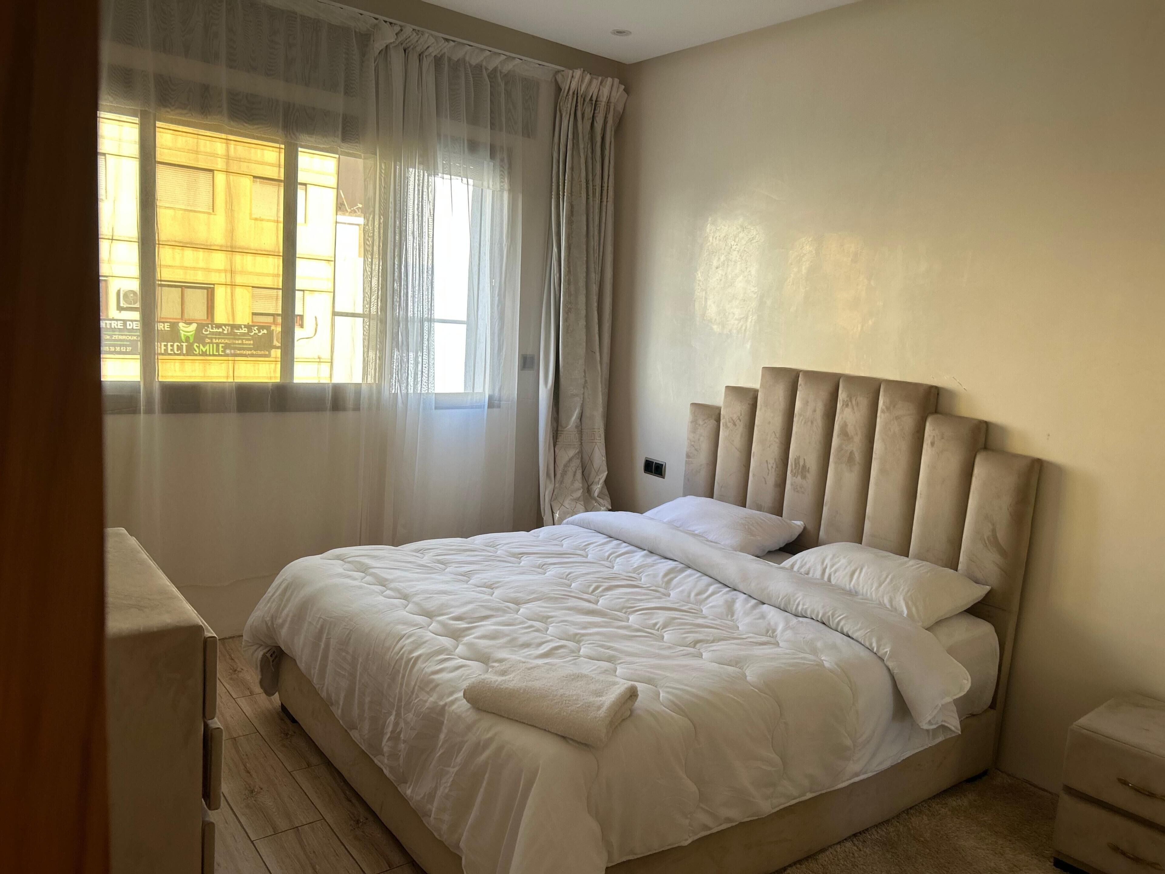 A bedroom with a large bed, a bedside table, a nightstand, and sheer curtains on a window.