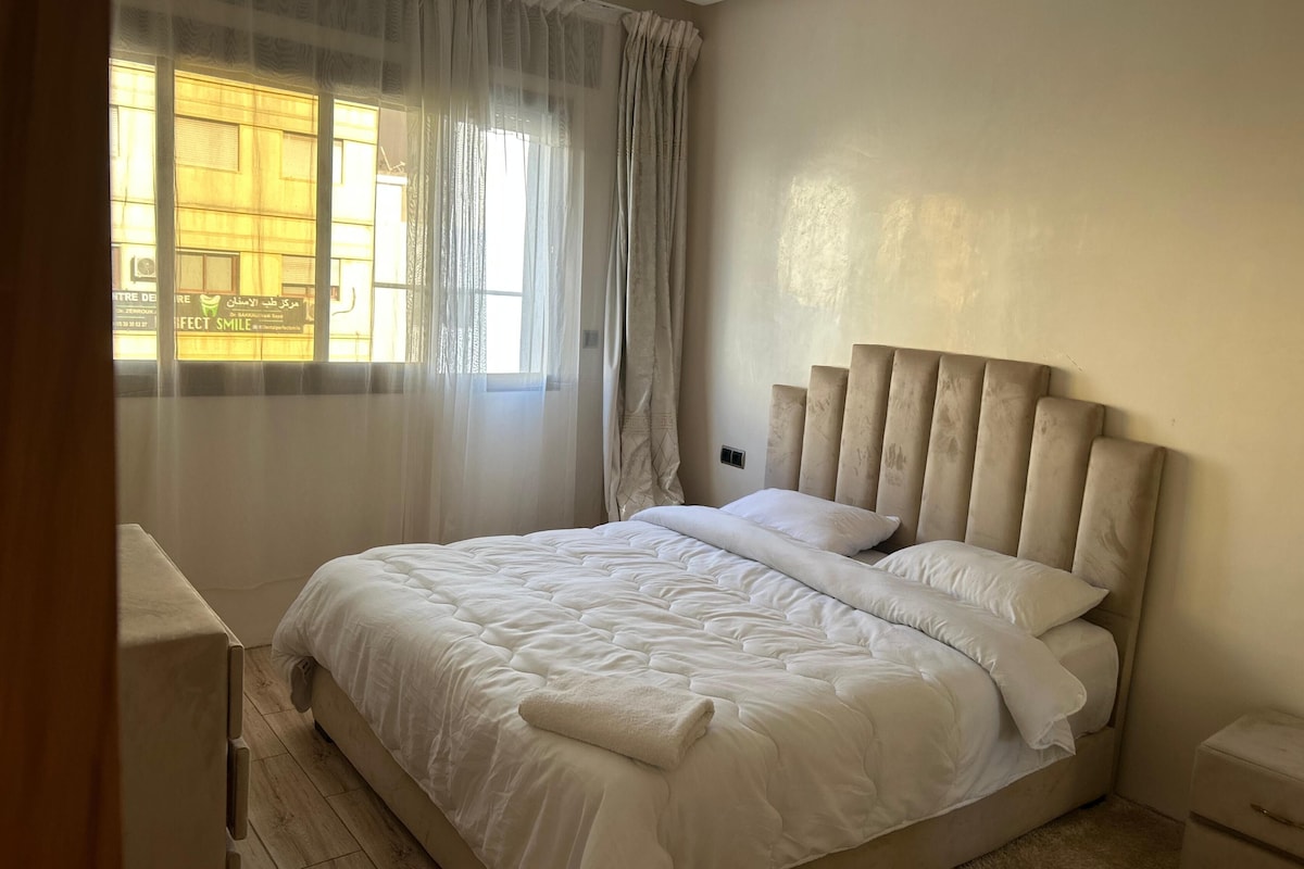A bedroom with a large bed, a bedside table, a nightstand, and sheer curtains on a window.