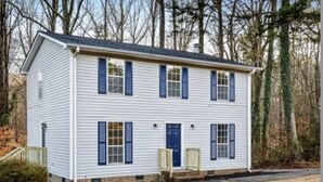 A two-story white house with blue shutters and a front door, surrounded by trees.