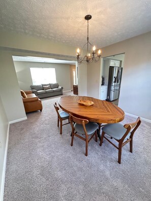 A dining area with a wooden table and chairs, a chandelier, and a view into a living room with a sofa and a kitchen area.