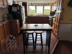 A kitchen with wooden cabinets, a central island with stools, a refrigerator, and a window with a view of greenery. - BOOK WITH US AT PARADISE ON THE GULF, BRING YOUR BOAT, DOCKAGE INCLUDED. (Hudson)