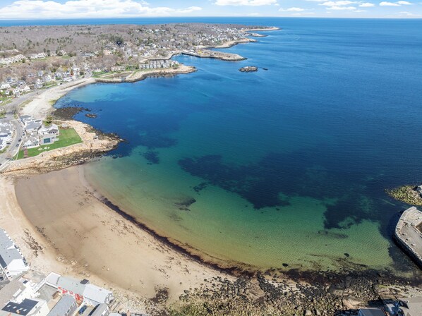 Vue aérienne d’une zone côtière avec une plage, des eaux cristallines et des immeubles résidentiels.