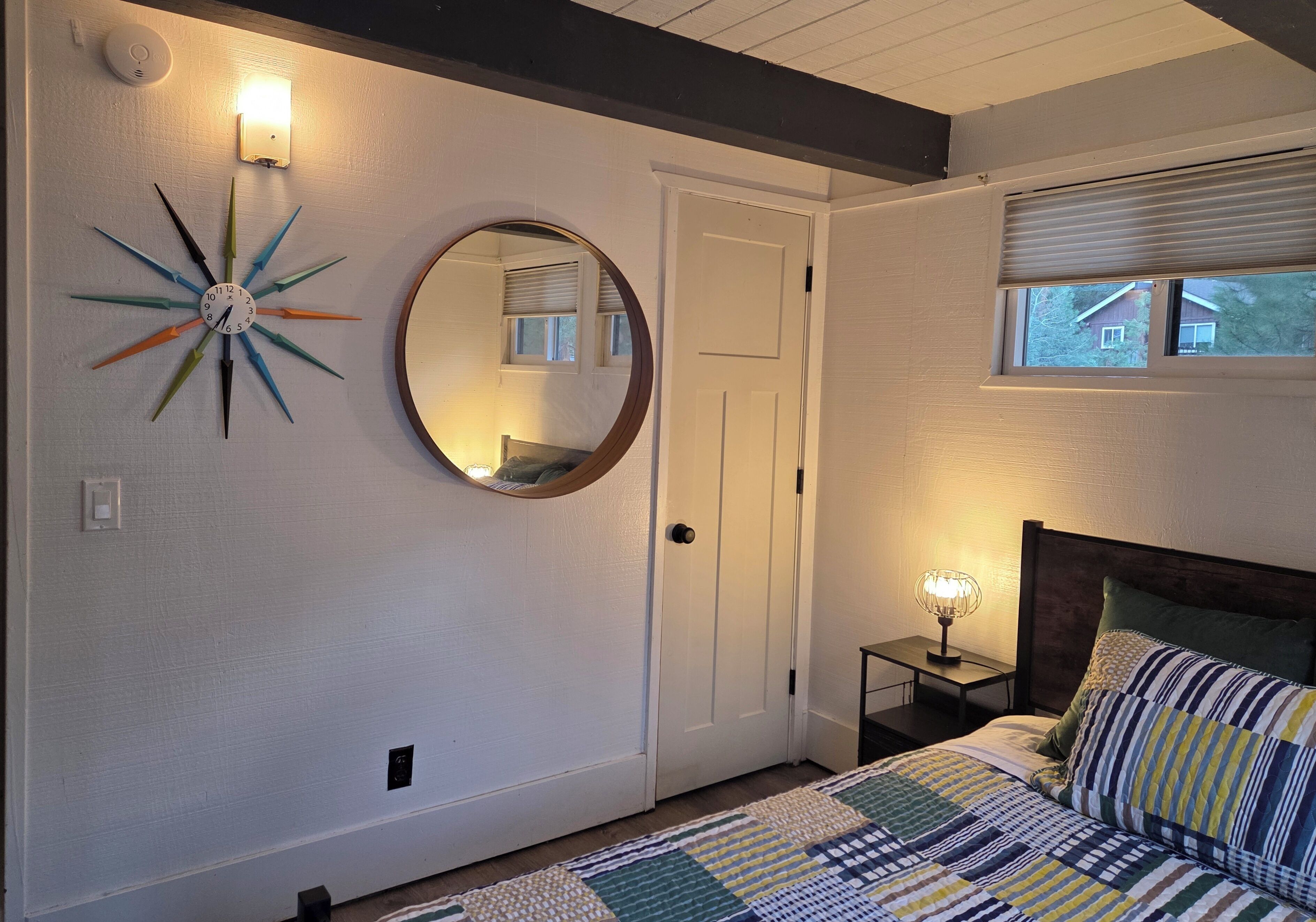 A bedroom with a bed, a round mirror, a wall clock, a bedside table with a lamp, and a window with blinds.