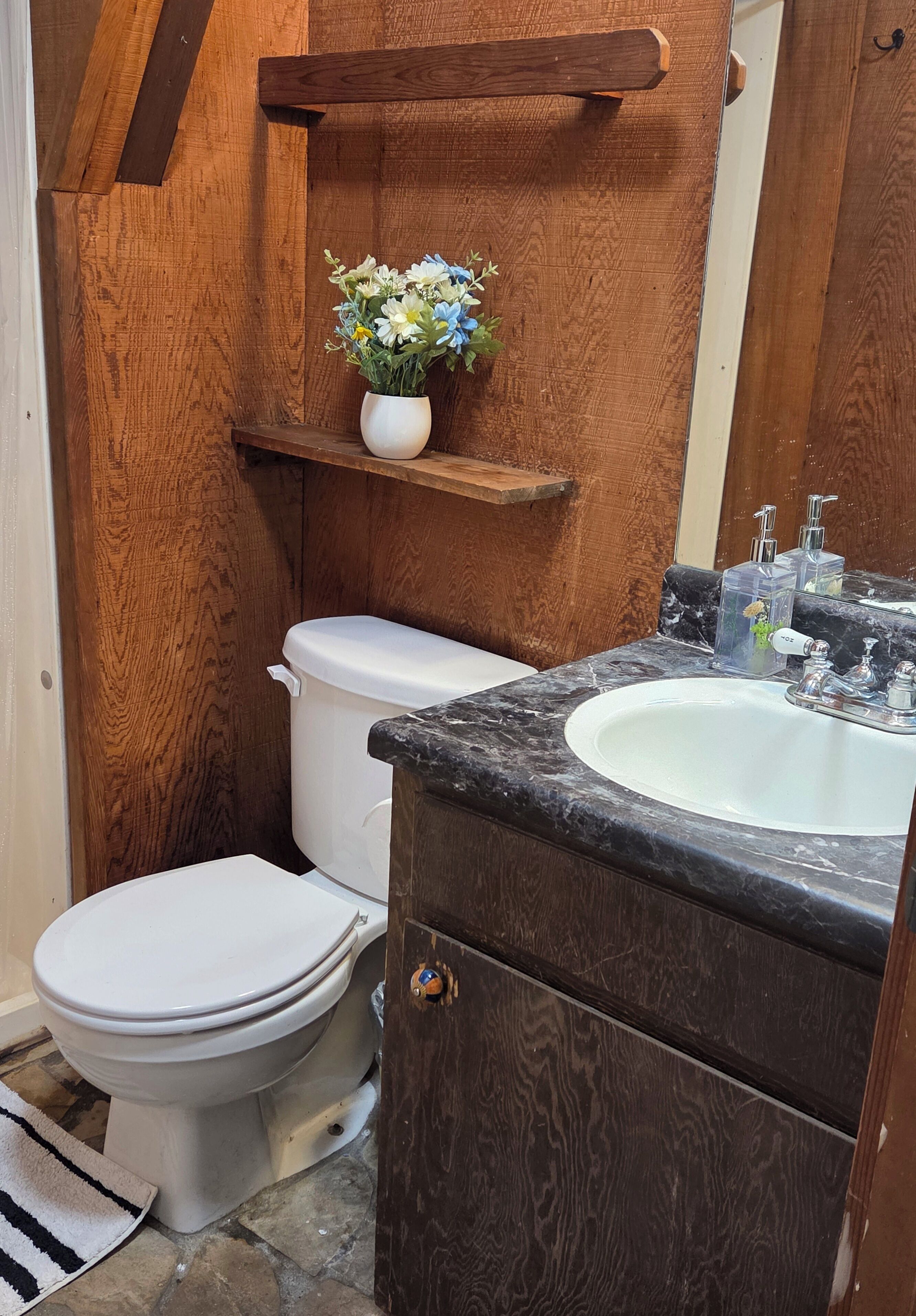 A compact bathroom with a white toilet, a marble sink, and a wooden shelf holding a vase of flowers.