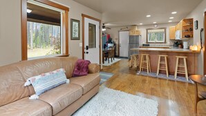 A cozy living room with a leather sofa, a white fluffy rug, and a kitchen area with wooden stools and a refrigerator. - New listing! Cozy 2 bed cabin 10 mins to Glacier (Coram)