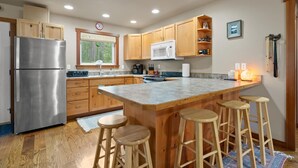 A modern kitchen with wooden cabinets, a stainless steel refrigerator, a microwave, and a large island with stools. - New listing! Cozy 2 bed cabin 10 mins to Glacier (Coram)
