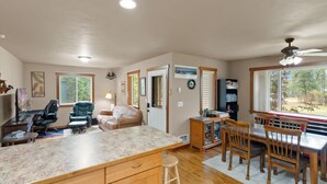 A spacious kitchen with a large island, wooden dining table, and chairs. There is a ceiling fan, a window with a view of greenery, and a door leading to another room. - New listing! Cozy 2 bed cabin 10 mins to Glacier (Coram)