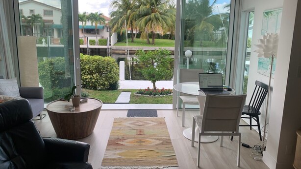 Een moderne woonkamer met een grote glazen deur die uitkomt op een balkon. De kamer heeft een zwarte leren bank, een ronde salontafel en een klein bijzettafeltje met een plant. Een laptop staat op een witte tafel, en er is een stoel met een zwarte rugleuning. Het balkon biedt uitzicht op een tuin met palmbomen en een gebouw op de achtergrond.