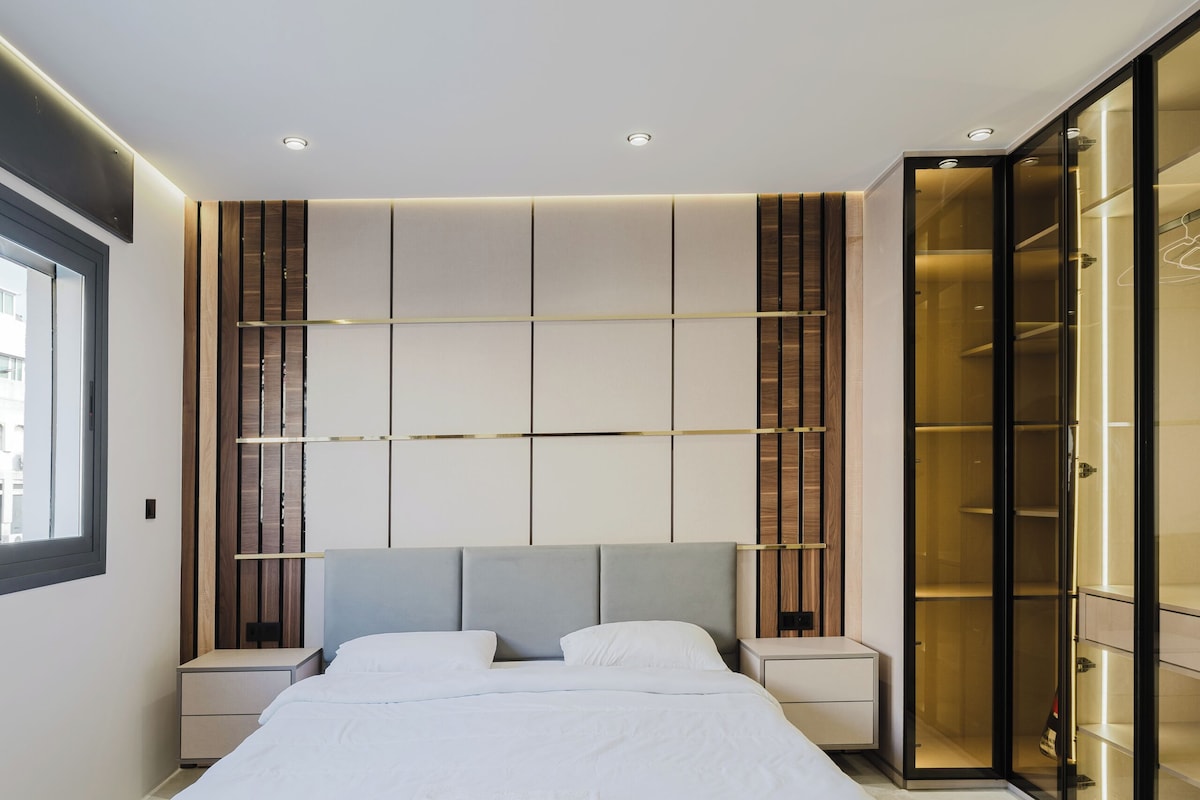 A modern bedroom with a large bed, bedside tables, a wooden bench, and a walk-in closet.