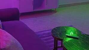 A room with a flat-screen TV mounted on the wall, a sofa, a small table, and a decorative hanging light fixture. - Serene Homes Membley- Executive 1-Bedroom in Ruiru- Secure & Affordable (Ruiru)