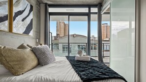 A modern hotel room with a large bed, a view of the cityscape through floor-to-ceiling windows, and a cityscape view from the balcony. - Luxury 2BR/2BA Condo in Calgary Beltline | King Bed, Free Underground Parking (Calgary)
