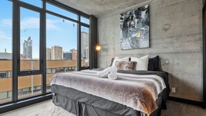 A modern bedroom with a large bed, a city view through floor-to-ceiling windows, and a concrete wall with a painting. - Luxury 2BR/2BA Condo in Calgary Beltline | King Bed, Free Underground Parking (Calgary)