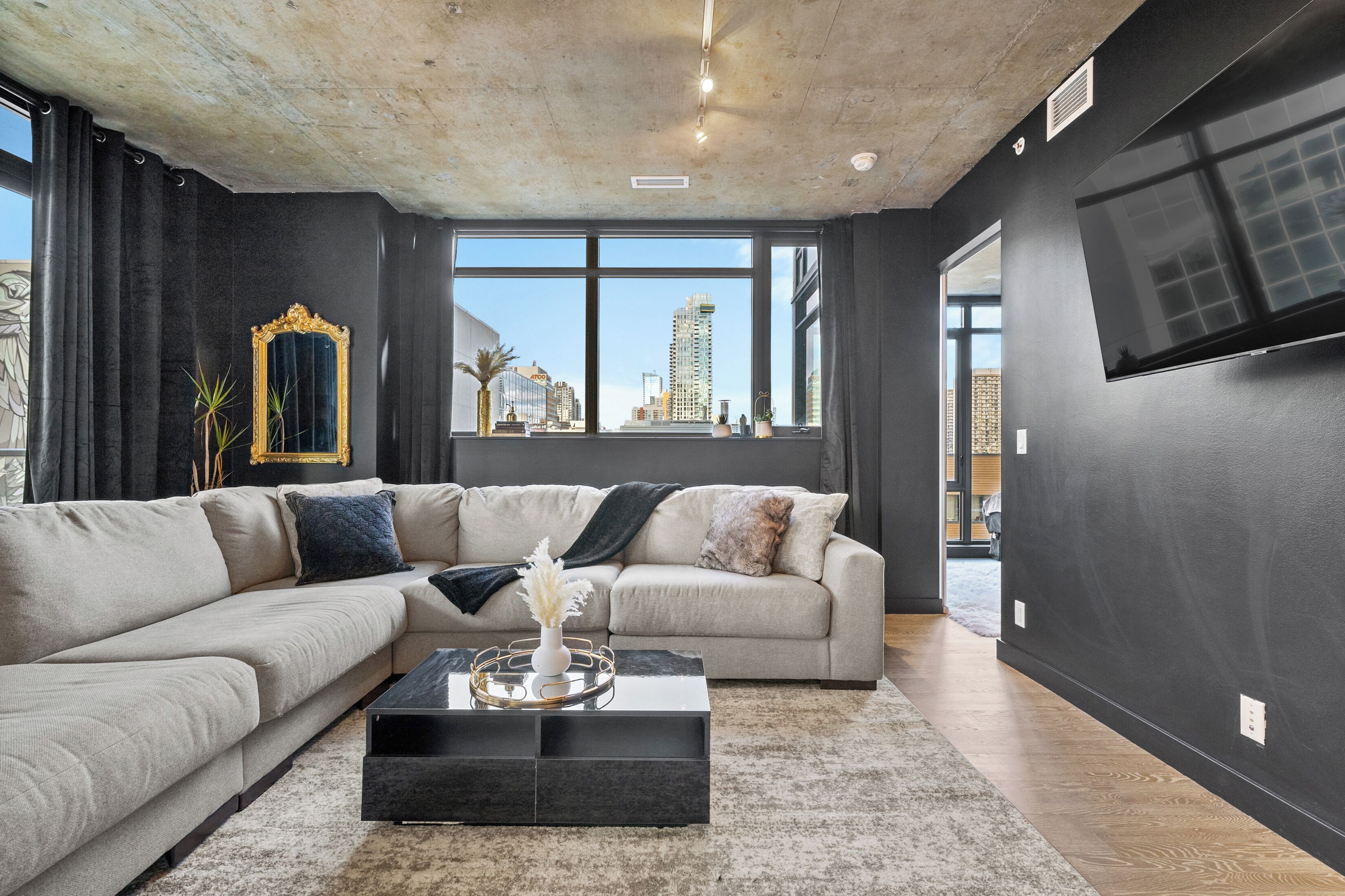 A modern living room with a sectional sofa, a coffee table with decorative items, a large window with a city view, and a flat-screen TV mounted on the wall.