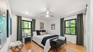 Une chambre à coucher comprenant un lit, des tables de chevet, un ventilateur de plafond et de grandes fenêtres avec des rideaux.