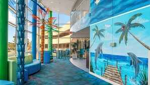 Amplo espaço interno com um mural colorido de golfinhos e palmeiras, áreas de estar e vista para o oceano através de grandes janelas.