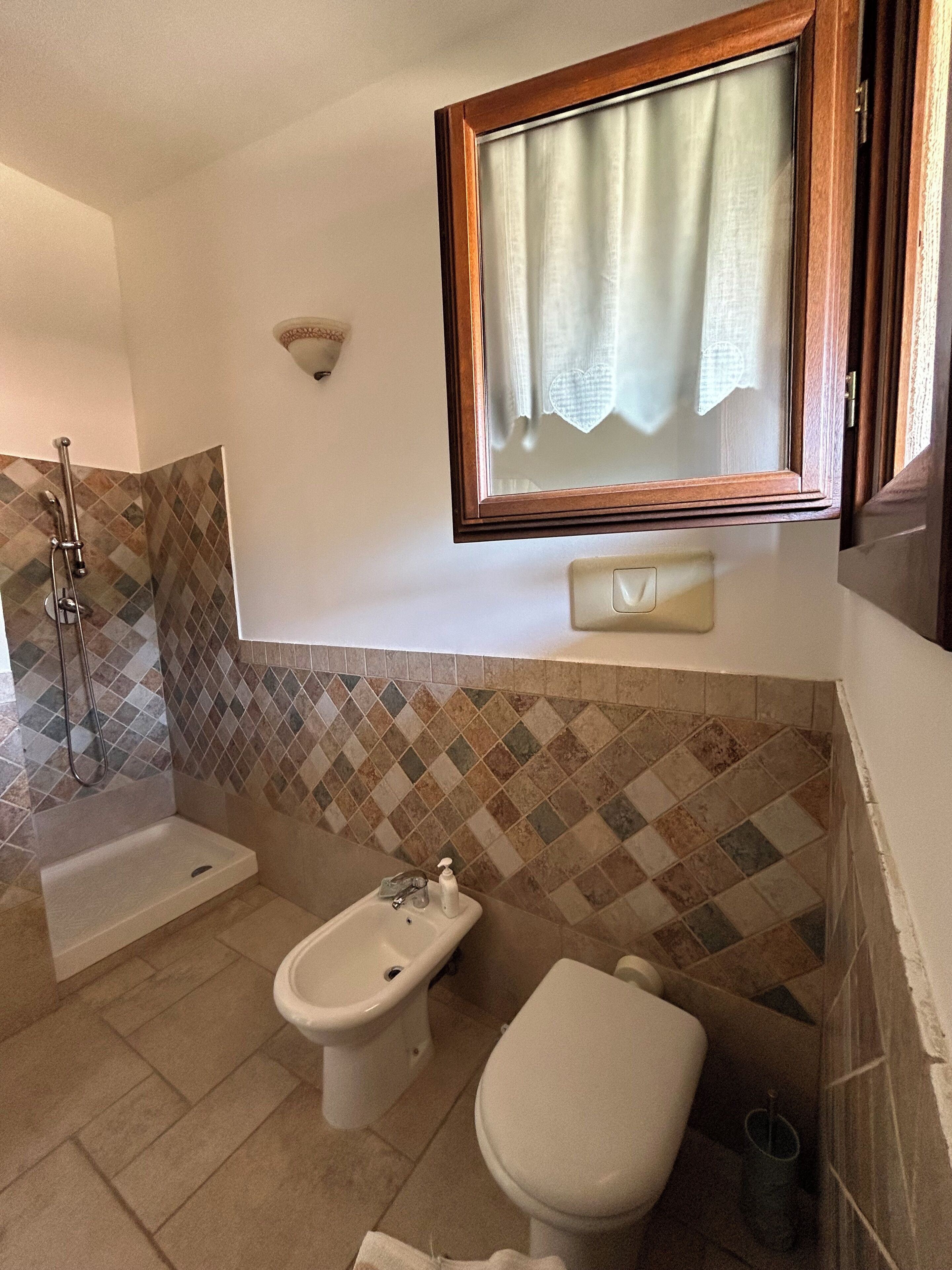 A bathroom with a shower, a bidet, a toilet, and a window with a curtain.