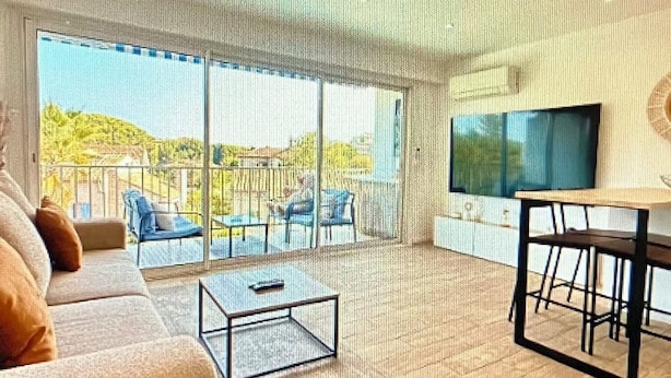 Uma sala de estar ampla, com uma grande porta de vidro deslizante que dá para uma varanda, um sofá, uma mesa de centro e uma televisão.